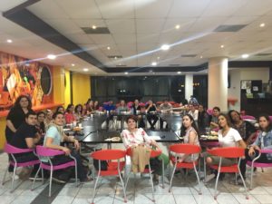 Large group of students sit around a rectangular cafeteria table smiling at the camera. 