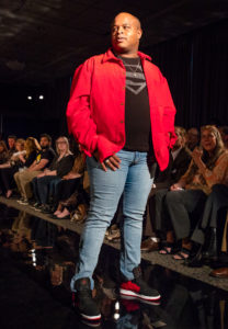 A male model poses at the end of the fashion runway looking in the distance with a moody expression. 
