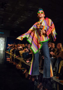A model in 1970's inspired disco wear poses at the end of the runway while holding a small dog in matching outfit. 