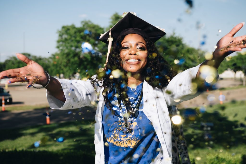 A woman stands with her hands held out, throwing glitter at the camera. She holds her head up high and wears a large smile, a graduation cap, and what appears to be a doctors jacket. 