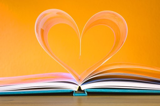 A teal hard covered book is laying with the pages open on a table, as if someone was reading, pictured with two pages folded up and together toward the  seam that looks like a heart. The backdrop is a bold yellow. 