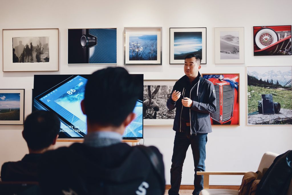 Background: A man stands in front of a wall almost covered in various pieces of visual art. He seems to be lecturing as he holds his hands in to his chest as if ready to throw them open, next to him appears to be a television. Foreground: Students sit with their head faced away from the camera, watching the man talk, we see the back of their heads.