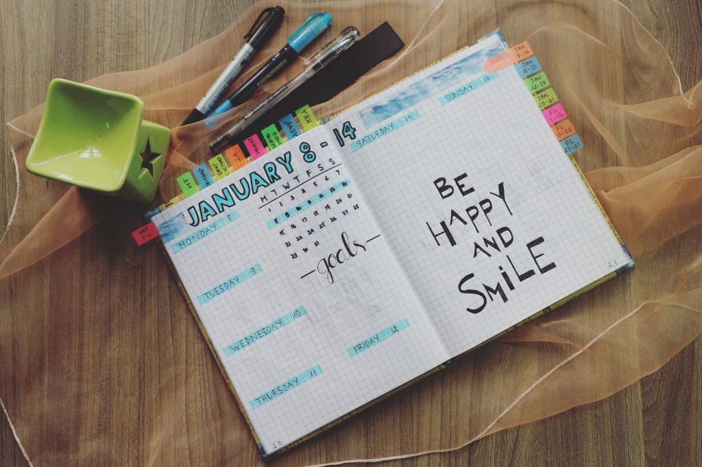 A weekly planner lays open in a table, the book lays atop a sheer piece of toole next to a scented wax melter and a handful of pens and markers. The book is littered with small posted notes and drawn on with a highlighter over all dates. The right page underneath the weekend says in bold "be happy and smile" 