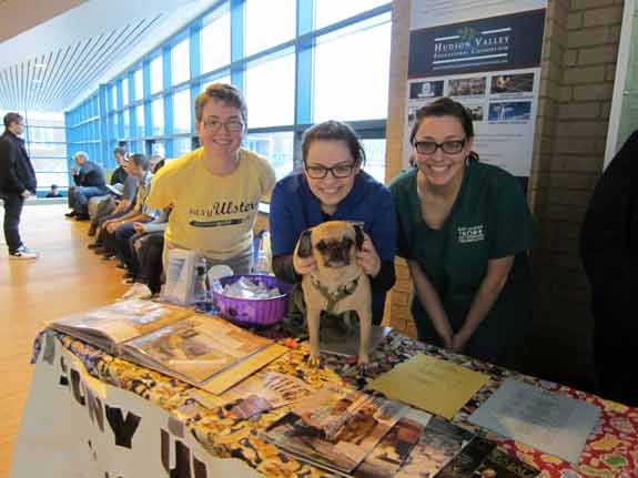 Three young women stand bowed over a table that has a dog sitting on top being held by the middle woman. The table is decorated and filled with veterinary information. Behind them is a hallway of windows with many people seated in front of them. 