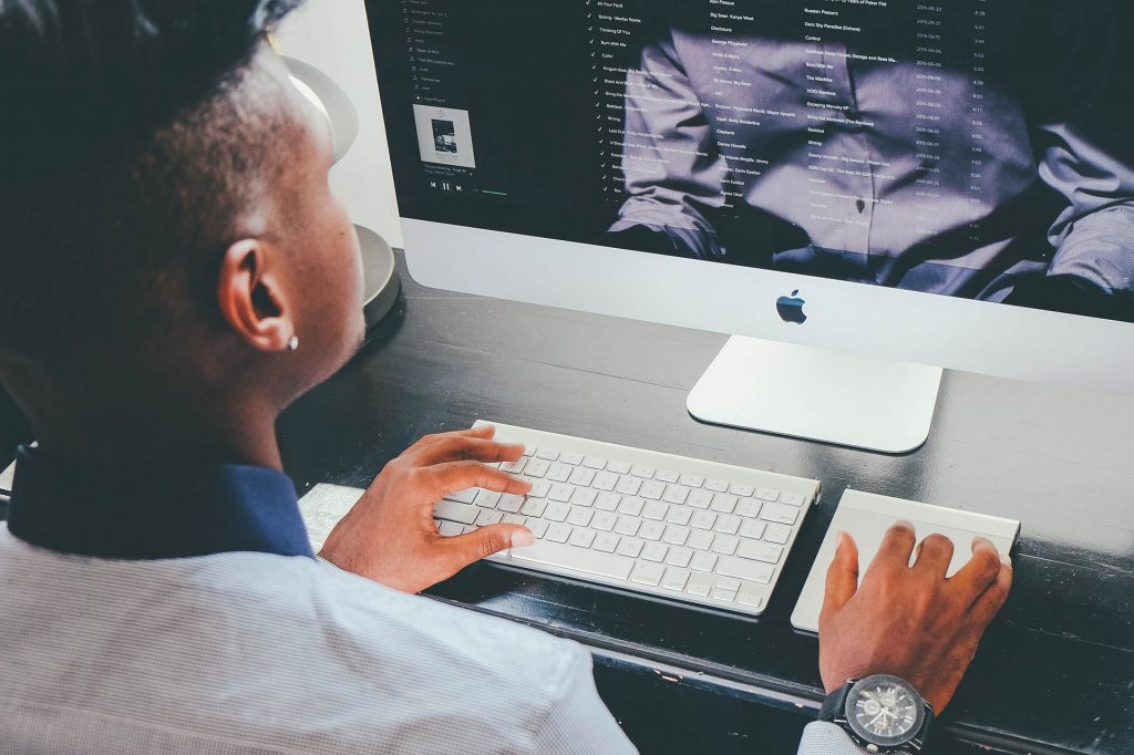 A dark skinned man sits at a mac computer and holds his hands on the mouse touch pad and keyboard. He is wearing a grey shirt with a dark blue collar, a watch, and has an earring in his right ear. We cannot see his face 