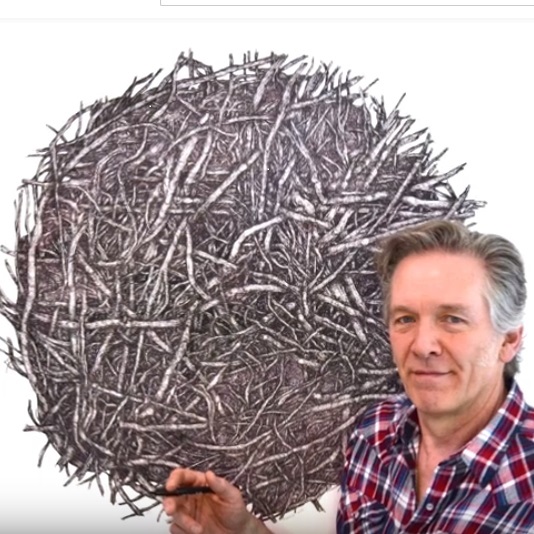 Artist Iain Machell stands in front of a pencil drawing of what appears to be a ball of hay or twigs. He is smiling and holding a pencil. 