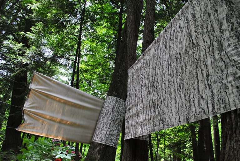 Two trees stand in middle of the frame and are wrapped with paper that features prints if their bark. The paper extends out of the frame, as if rolling off the tree.