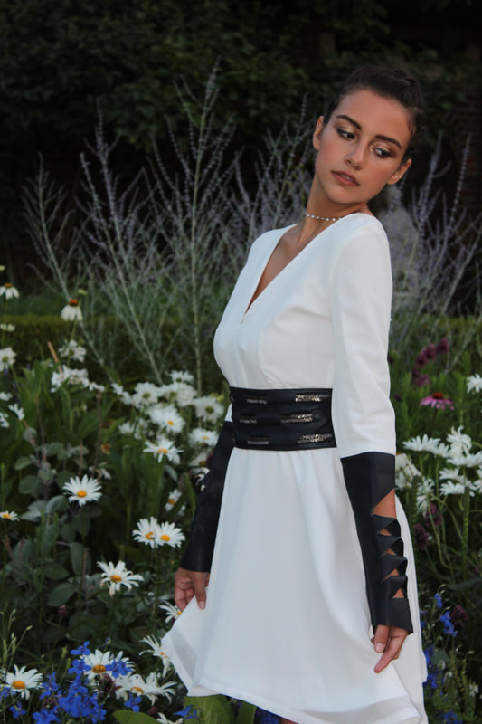 A woman stands in a white dress with leather sleeve and belt accents
