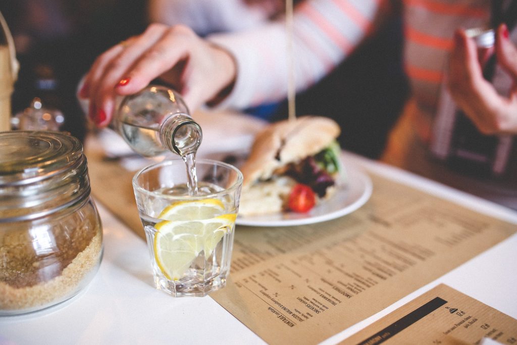 A hand with painted nails reached over a glass and is pouring water out of a glass bottle into the glass on the table.