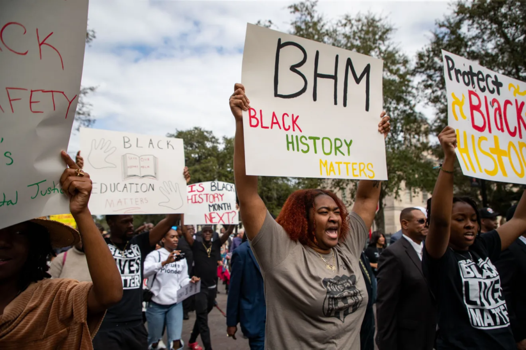 Black Americans hold signs saying "Black History Matters" and "Protect Black History" while walking down the street