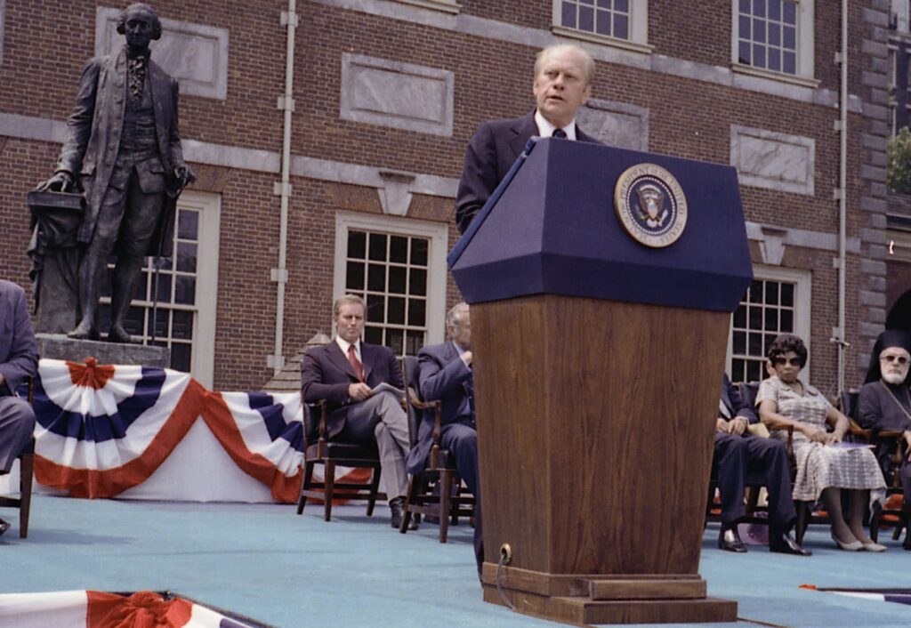 President Ford stands at a podium on a stage addressing a crowd