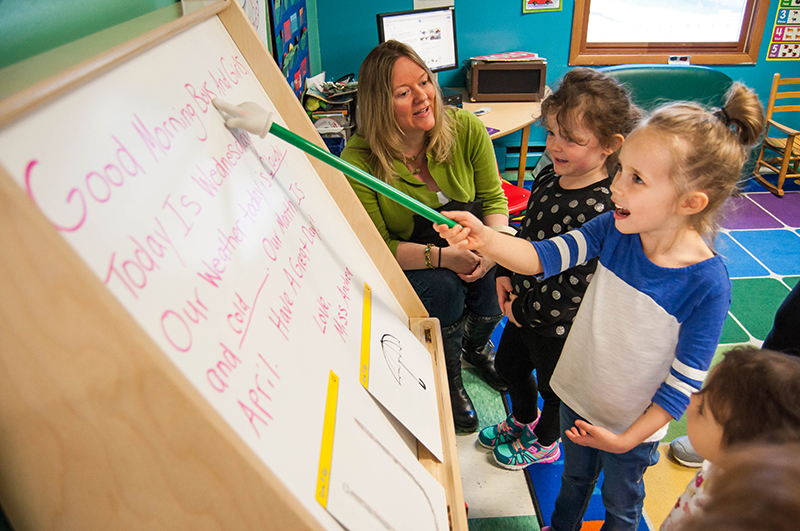 Child pointing at board while teacher looks on