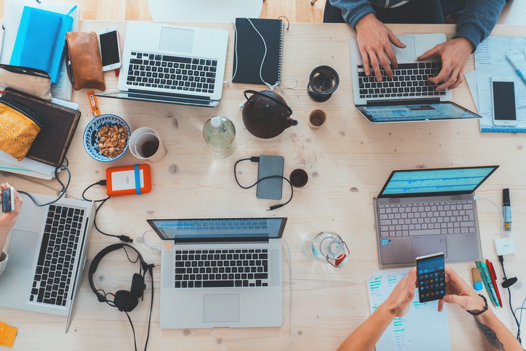overhead view of work table covered in laptops, tea cups, smartphones, pens, cords, and some hands typing