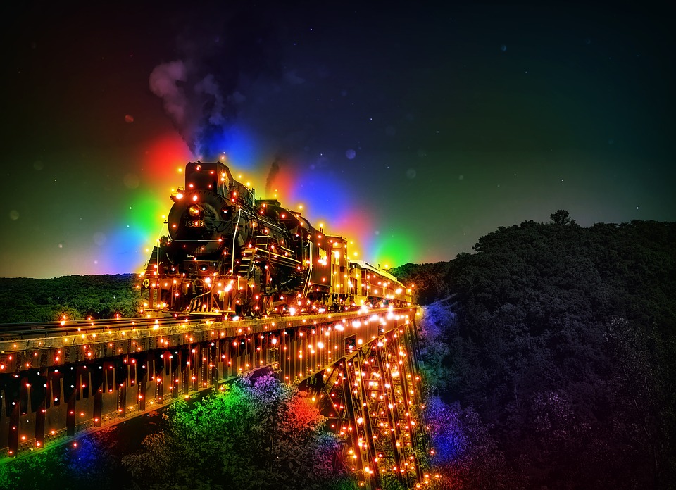 A dark forest landscape with set of train tracks leading out of the forest and off the side of the photo, the tracks and the train on top of them are covered and lit completely by rainbow christmas lights