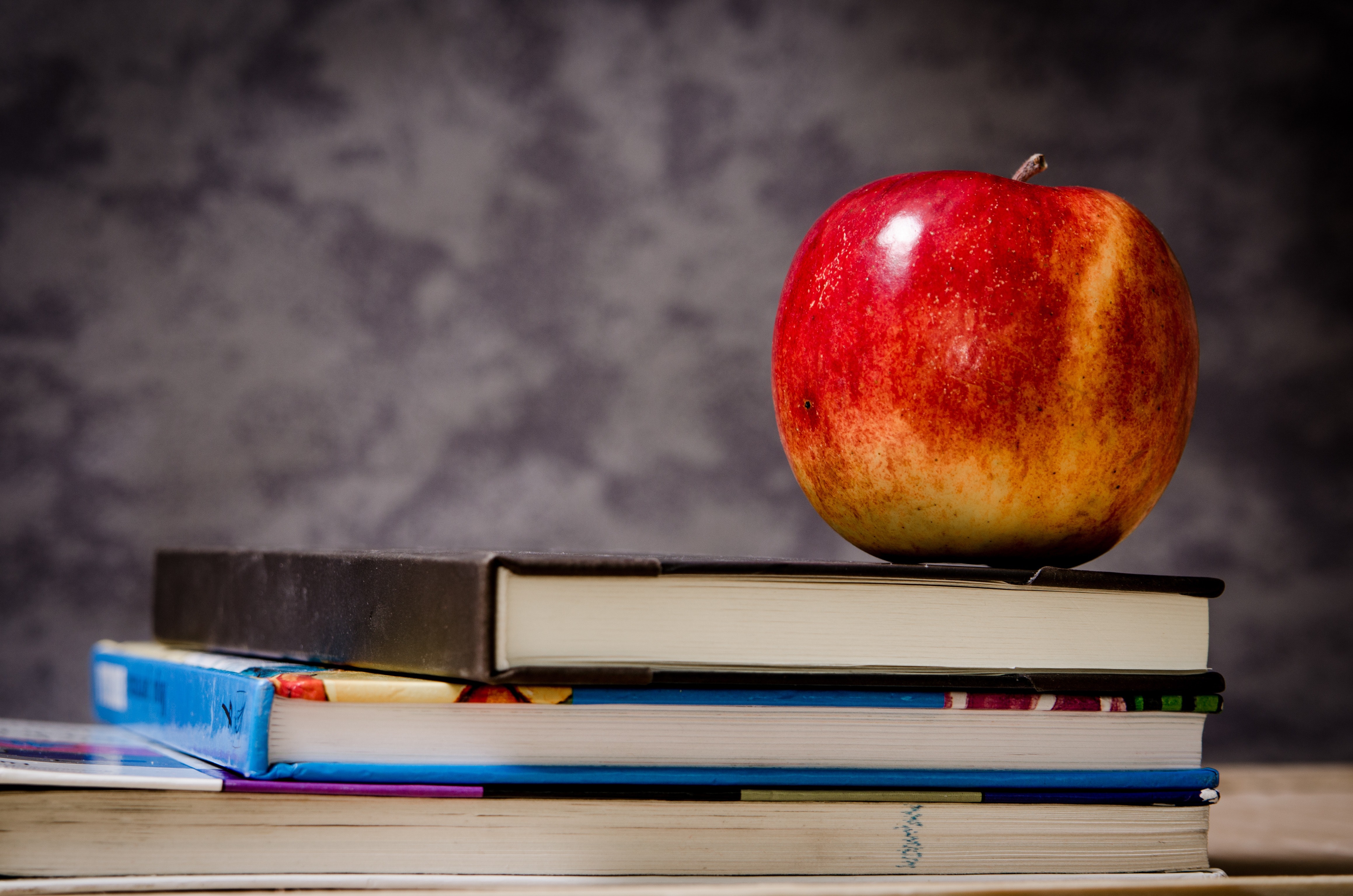 A bright red apple sits atop a stack of three hard cover books, we cannot see their titles or covers. This display sits atop a table in front of a smoky grey background.