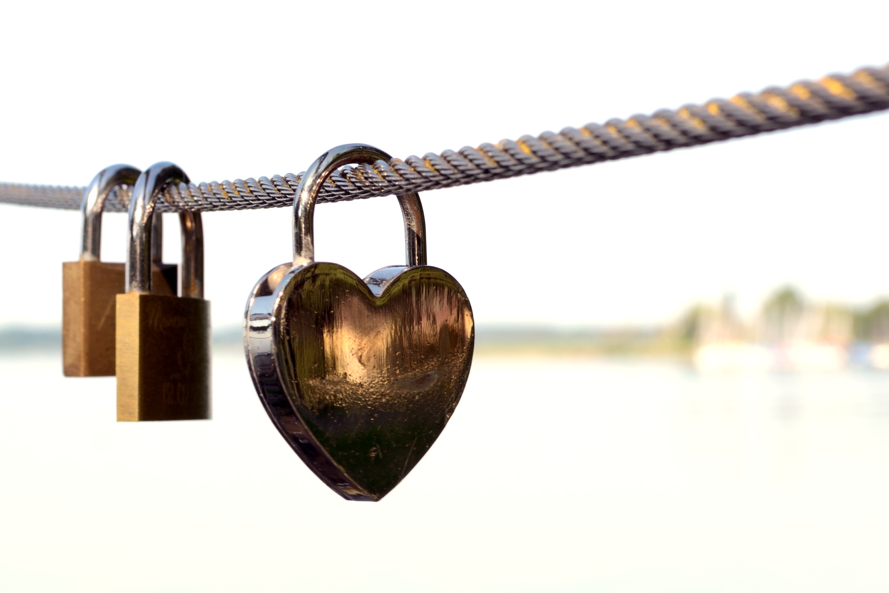 A very light washed out background is shown with a rope hanging in the foreground. The rope has three locks hooked to it. Two are smaller square locks and one is larger heart shaped lock closer to the camera than the other smaller locks.