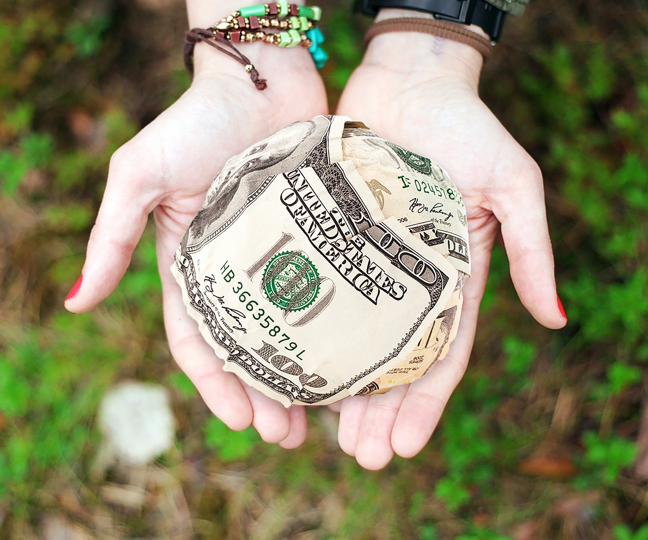 Two open faced hands hold a big ball of crumpled bills in them above a brown and green background of blurry foliage. A metaphoric representation of financial aid such as FAFSA