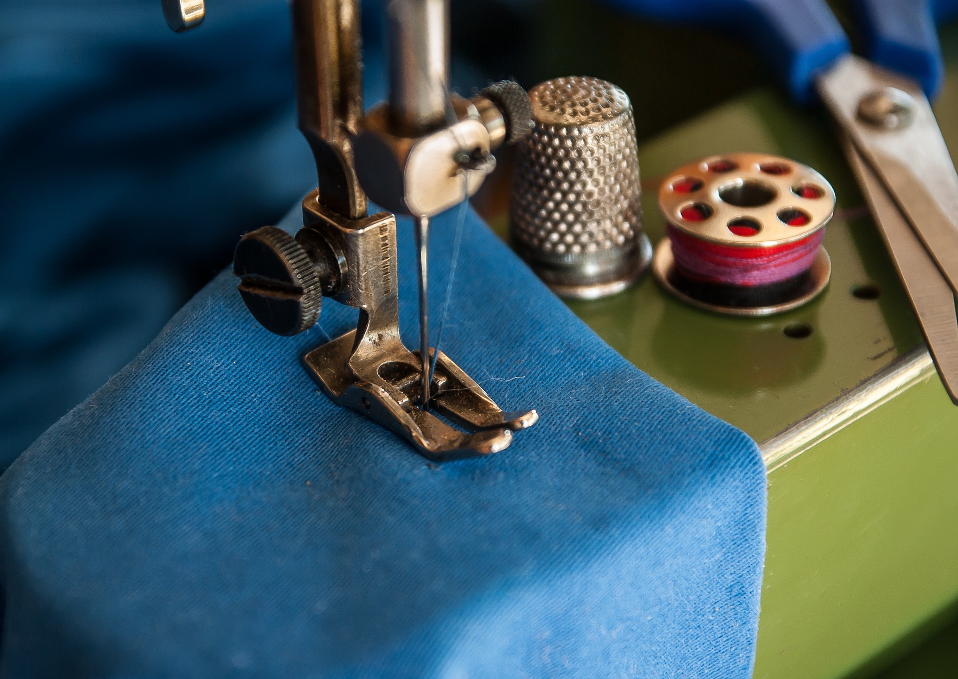 A green sewing machine works on a blue cloth with a spool of red thread sitting next to a pair of scissors