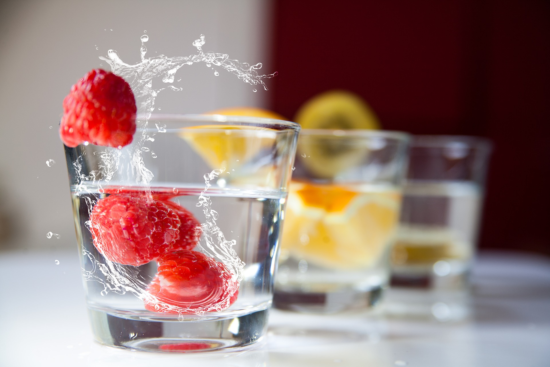 Three glasses of water sit on a table with fruit sitting in them, the closest glass has a raspberry splashing into the water in the glass.