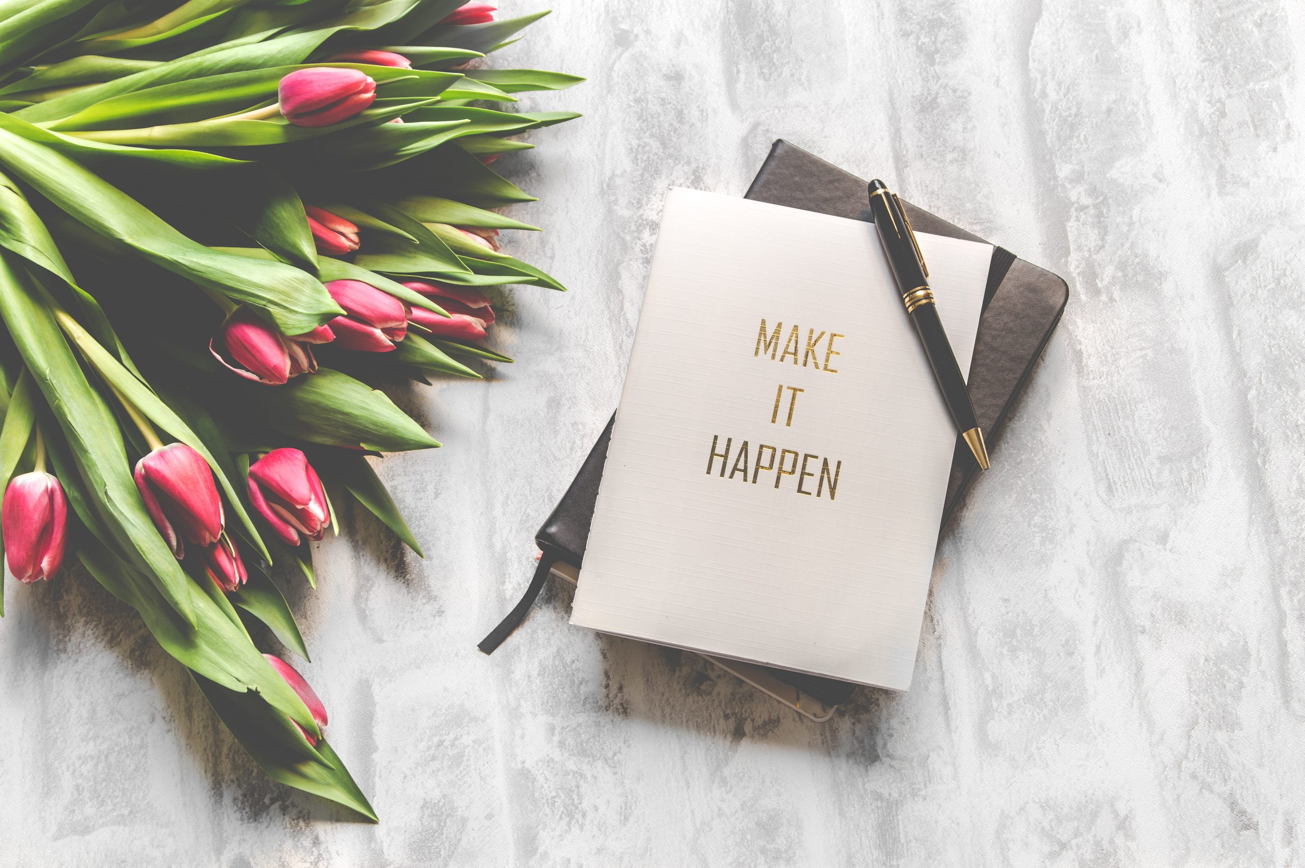 A bouquet of flowers sits in the corner of the image of a table top from an aerial view. Also on the table is two notebooks, one of which says, "make it happen".
