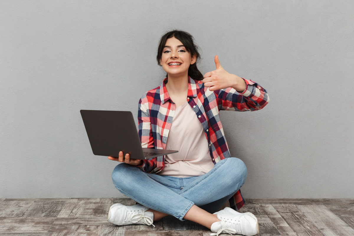 Student sits cross-legged while holding a laptop in one hand and giving a thumbs up with the other